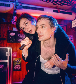 Zwei Personen auf der Bühne in einem Pub. Sie singen betrunken Karaoke. Sie sitzt auf seinem Rücken.