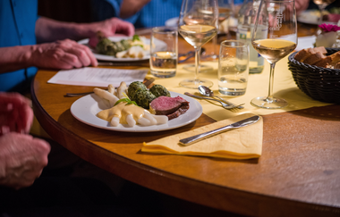 Holztisch, eingedeckt mit gelber Tischdecke ; Teller mit Spargel, Braten und Spinatknödeln | © Christian Seyffer