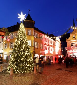 Ein festlich geschmückter und hell erleuchteter Weihnachtsbaum steht inmitten eines Platzes, im Hintergrund befinden sich beleuchtete Häuser