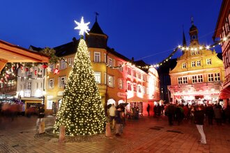Ein festlich geschmückter und hell erleuchteter Weihnachtsbaum steht inmitten eines Platzes, im Hintergrund befinden sich beleuchtete Häuser
