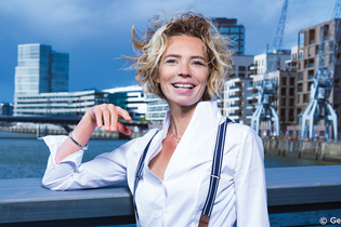 Frau mit lockigem Haar und weißem Hemd steht lächelnd vor einer modernen Stadtlandschaft mit Wasser und Gebäuden. | © Kultur- und Sportamt Stadt Bietigheim-Bissingen
