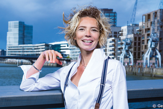 Frau mit lockigem Haar und weißem Hemd steht lächelnd vor einer modernen Stadtlandschaft mit Wasser und Gebäuden. | © Kultur- und Sportamt Stadt Bietigheim-Bissingen