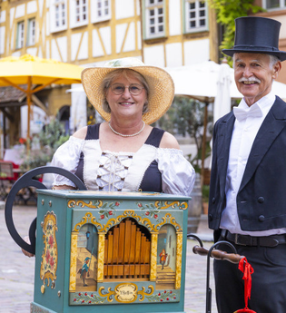 Ein Mann und eine Frau in historischer Kleidung stehen neben einer dekorativen Drehorgel auf einem gepflasterten Platz mit Fachwerkhäusern im Hintergrund. | © Stadt Besigheim