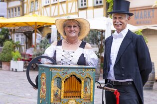 Ein Mann und eine Frau in historischer Kleidung stehen neben einer dekorativen Drehorgel auf einem gepflasterten Platz mit Fachwerkhäusern im Hintergrund. | © Stadt Besigheim
