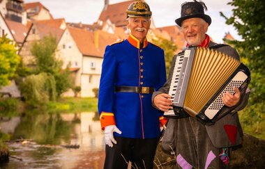 Zwei Männer in historischen Kostümen, einer in einer blauen Uniform und der andere mit einem Akkordeon, stehen vor einem Fluss in einer malerischen Stadt. | © Stadt Besigheim