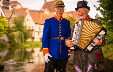 Zwei Männer in historischen Kostümen, einer in einer blauen Uniform und der andere mit einem Akkordeon, stehen vor einem Fluss in einer malerischen Stadt. | © Stadt Besigheim