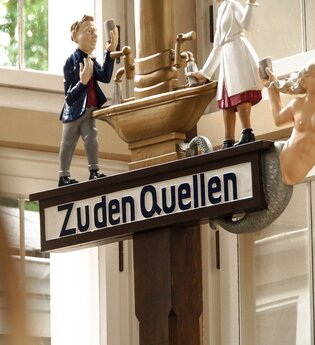Ein Schild mit der Aufschrift "Zu den Quellen" zeigt drei Figuren: einen Mann in Anzug, eine Dame die das Heilwasser ausschenkt und eine Meerjungfrau. | © Kurverwaltung Bad Mergentheim