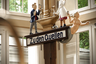 Ein Schild mit der Aufschrift "Zu den Quellen" zeigt drei Figuren: einen Mann in Anzug, eine Dame die das Heilwasser ausschenkt und eine Meerjungfrau. | © Kurverwaltung Bad Mergentheim