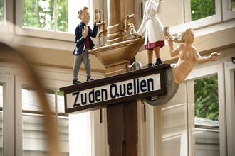 Ein Schild mit der Aufschrift "Zu den Quellen" zeigt drei Figuren: einen Mann in Anzug, eine Dame die das Heilwasser ausschenkt und eine Meerjungfrau. | © Kurverwaltung Bad Mergentheim