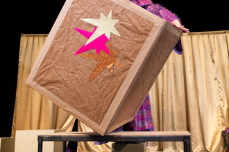 Künstlerin auf der Bühne mit einem lebensgroßen Kiste vor sich balancierendLLY - Familientheater