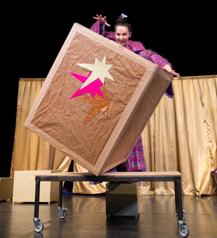 Künstlerin auf der Bühne mit einem lebensgroßen Kiste vor sich balancierendLLY - Familientheater