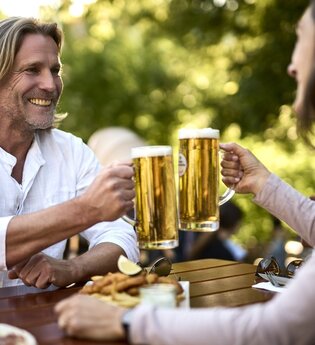 Eine Frau und ein Mann stoßen mit zwei Gläsern Bier an. Sie sitzen an einem Holztisch in der Natur. Vor ihnen steht etwas zum Essen. | © Tourimia Tourismus GmbH | Stefan Leitner Photography