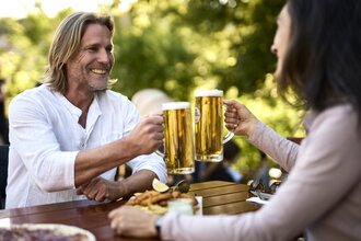 Eine Frau und ein Mann stoßen mit zwei Gläsern Bier an. Sie sitzen an einem Holztisch in der Natur. Vor ihnen steht etwas zum Essen. | © Tourimia Tourismus GmbH | Stefan Leitner Photography