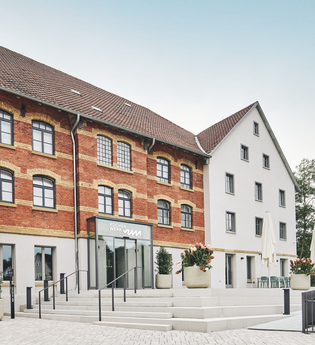 Zwei Gebäude, eines aus rotem Backstein und eines weiß, mit einem Eingang und Treppen. Blumenkübel und Tische mit Sonnenschirmen sind vorhanden. | © Kocherwerk