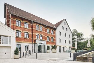 Zwei Gebäude, eines aus rotem Backstein und eines weiß, mit einem Eingang und Treppen. Blumenkübel und Tische mit Sonnenschirmen sind vorhanden.