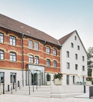 Zwei Gebäude, eines aus rotem Backstein und eines weiß, mit einem Eingang und Treppen. Blumenkübel und Tische mit Sonnenschirmen sind vorhanden.