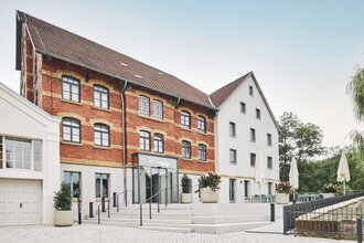Zwei Gebäude, eines aus rotem Backstein und eines weiß, mit einem Eingang und Treppen. Blumenkübel und Tische mit Sonnenschirmen sind vorhanden.