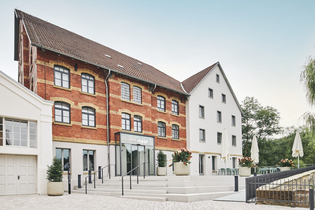 Zwei Gebäude, eines aus rotem Backstein und eines weiß, mit einem Eingang und Treppen. Blumenkübel und Tische mit Sonnenschirmen sind vorhanden. | © Kocherwerk