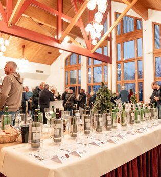 Auf einem Tisch steht eine Auswahl an Fürstenfass Weinen mit Informationen zu jeder Sorte. im Hintergrund stehen Menschen, die Wein trinken und sich unterhalten. | © Touristikgemeinschaft Hohenlohe e. V. | Samuel Eisel