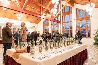 Auf einem Tisch steht eine Auswahl an Fürstenfass Weinen mit Informationen zu jeder Sorte. im Hintergrund stehen Menschen, die Wein trinken und sich unterhalten. | © Touristikgemeinschaft Hohenlohe e. V. | Samuel Eisel