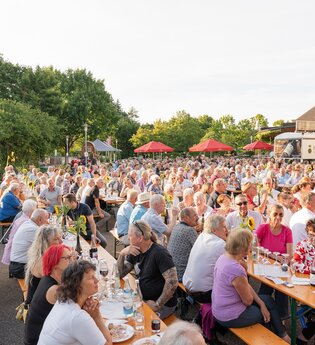 Viele Menschen sitzen auf Sitzbänken an Tischen beim FUERSTENFASS Sommerweinfest. | © Touristikgemeinschaft Hohenlohe e. V. | Samuel Eisel
