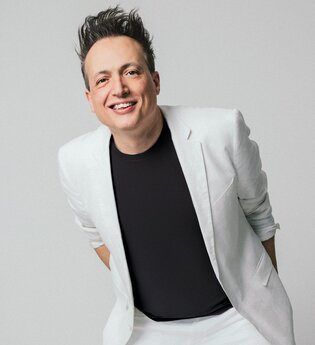 Der Comedian und Musiker Olaf Bossi in weißen Sakko und schwarzem T-Shirt steht lächelnd vor einem grauen Hintergrund.