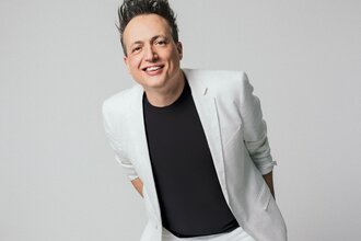 Der Comedian und Musiker Olaf Bossi in weißen Sakko und schwarzem T-Shirt steht lächelnd vor einem grauen Hintergrund.