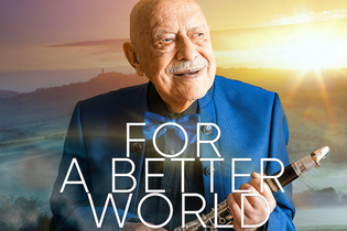 Ein älterer Mann in einem blauen Anzug hält eine Klarinette vor einer Landschaft bei Sonnenuntergang. Text: Maestro Giora Feidman - For a Better World. | © MACC Management GmbH