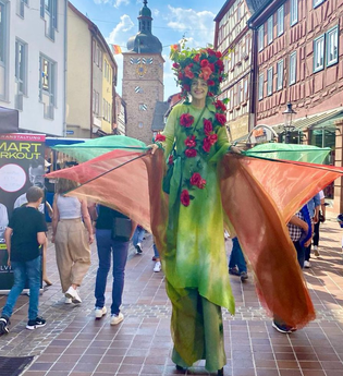 Eine Person auf Stelzen trägt ein grünes Kostüm, geschmückt mit roten Rosen, und bewegt sich durch die Fußgängerzone von Buchen. Im Hintergrund ragt ein Turm empor. | © Stadt Buchen