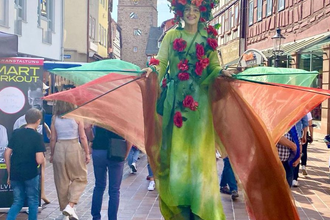 Eine Person auf Stelzen trägt ein grünes Kostüm, geschmückt mit roten Rosen, und bewegt sich durch die Fußgängerzone von Buchen. Im Hintergrund ragt ein Turm empor. | © Stadt Buchen
