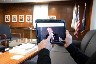 Im Vordergrund ein Tablet, das von einem Mann gehalten wird. Im Hintergrund ein großer Schreibtisch im Kommandantenzimmer des Mark Twain Centers. | © Kommandantenzimmer im Mark Twain Center. Foto: Christian Buck
