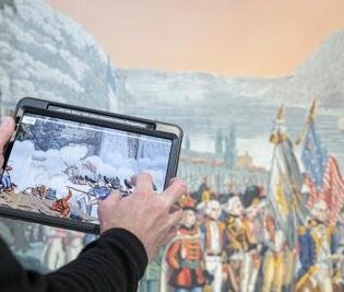 Im Vordergrund ein Tablet, das von einem Mann gehalten wird. Im Hintergrund eine historische Tapete.