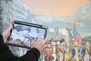 Im Vordergrund ein Tablet, das von einem Mann gehalten wird. Im Hintergrund eine historische Tapete.