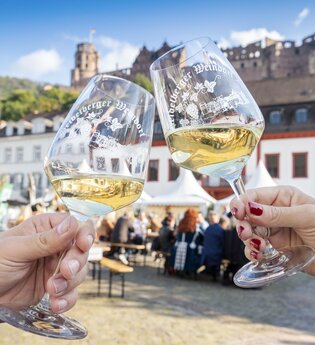 Zwei Personen stoßen mit Weingläsern auf dem Heidelberger Weindorf an, im Hintergrund der Karlsplatz und das Heidelberger Schloss. | © Heidelberg Marketing GmbH / Tobias Schwerdt
