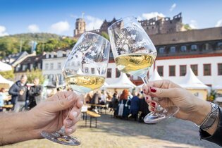 Zwei Personen stoßen mit Weingläsern auf dem Heidelberger Weindorf an, im Hintergrund der Karlsplatz und das Heidelberger Schloss. | © Heidelberg Marketing GmbH / Tobias Schwerdt