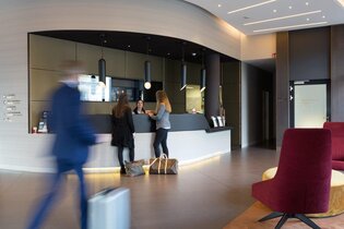 Im Bildhintergrund ist die Rezeption eines Hotels zu sehen. Zwei Frauen sind im Gespräch mit einer Mitarbeiterin an der Rezeption. Reisetaschen stehen am Boden. Ein Mann im blauen Anzug mit silbernem Trolly läuft auf die Rezeption zu