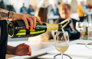 Sekt wird in ein Glas eingeschenk, im Hintergrund Gäste auf einem Weinfest