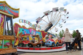 Auf dem Bild ist ein bunter Vergnügungspark mit einem Kinderkarussell, und einem Riesenrad im Hintergrund zu sehen. Mehrere Erwachsene und Kinder stehen davor oder gehen daran vorbei, während die farbenfrohen Fahrgeschäfte eine fröhliche Volksfestatmosphä | © Gemeinde Blaufelden