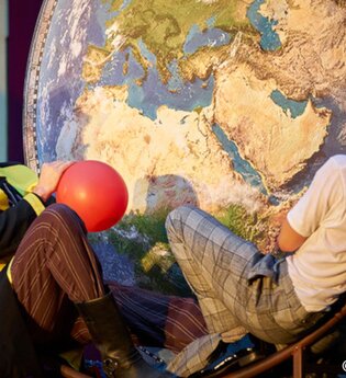 Zwei Schauspieler vor einer großen Weltkarte, einer hält einen roten Ballon. | © Kultur- und Sportamt Stadt Bietigheim-Bissingen
