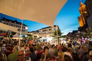 Gut mit Besuchern gefüllter Kiliansplatz in Abendstimmung. Die Kilianskirche ist beleuchtet und die Sonnensegel sind zu sehen.