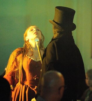 Schauspielszene mit Frau in rotem Kleid und Mann in schwarzem Mantel und Zylinder. | © Gruseldinner GmbH & Co. KG