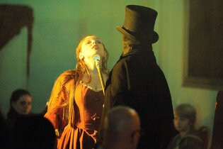 Schauspielszene mit Frau in rotem Kleid und Mann in schwarzem Mantel und Zylinder. | © Gruseldinner GmbH & Co. KG