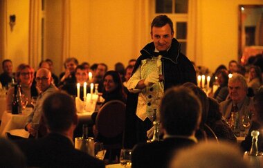 Ein Mann in einem Kostüm spricht zu einem Publikum in einem Kerzenlicht-Dinner. | © Gruseldinner GmbH & Co. KG