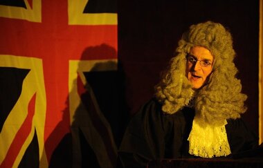 Ein Mann in historischer Richterrobe vor einer britischen Flagge. | © Gruseldinner GmbH & Co. KG