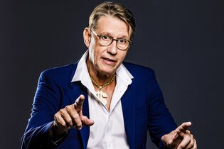 Ein Mann in einem blauen Anzug und weißem Hemd gestikuliert vor einem dunklen Hintergrund. | © Konzertkreis Triangel Compagnia Compostelli e.V.