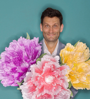 Ein Mann hält drei große, bunte Blumen vor einem türkisfarbenen Hintergrund. | © Konzertkreis Triangel Compagnia Compostelli e.V.