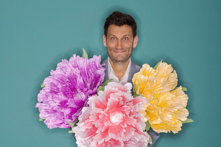 Ein Mann hält drei große, bunte Blumen vor einem türkisfarbenen Hintergrund. | © Konzertkreis Triangel Compagnia Compostelli e.V.