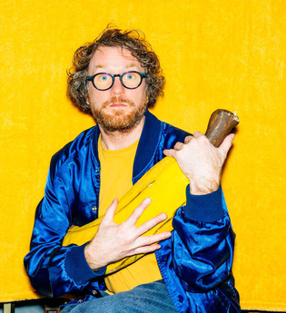 Ein Mann mit Brille und Bart hält ein großes gelbes Objekt vor einem gelben Hintergrund. | © Konzertkreis Triangel Compagnia Compostelli e.V.