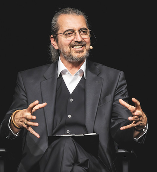 Ein Mann in einem Anzug sitzt auf einer Bühne und spricht in ein Mikrofon. | © Konzertkreis Triangel Compagnia Compostelli e.V.