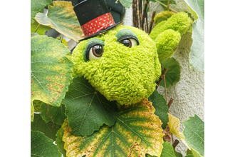 Ein grünes Plüschtier mit Zylinder schaut aus einem Blätterbusch hervor. | © Stadt Besigheim
